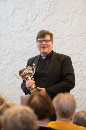 Antilla on käsissään komea pokaali ja hän hymyilee. Edessä istuu ihmisiä.