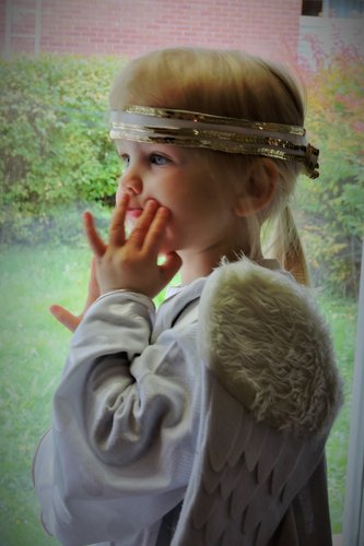 Pieni tyttö päällään enkelipuku ja -siivet katselee ikkunasta ulos.