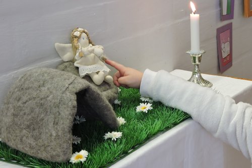 Enkeli istuu kiven päällä tyhjän haudan vieressä alttaripöydällä, jossa palaa kynttilä.