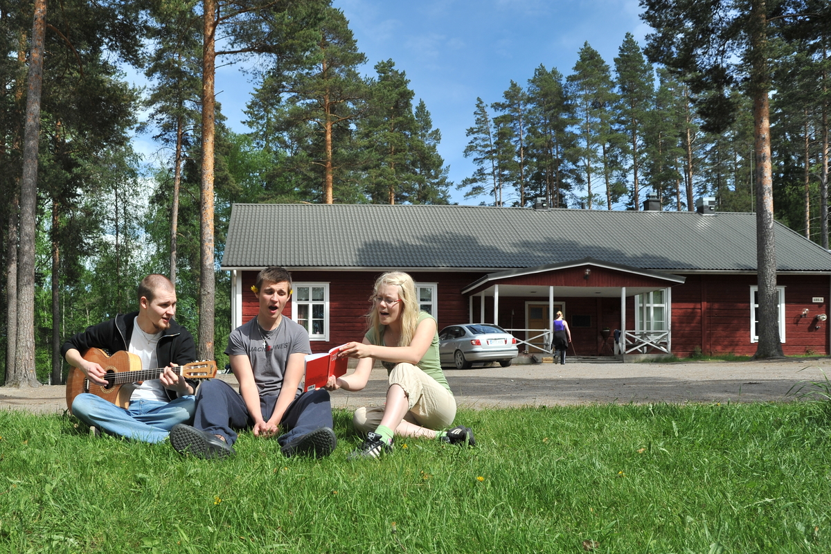 Lummeniemen leirikeskus Kesäinen kuva, Lummeniemen punainen hirsirakennus taustalla, edessä kolme nuorta laulaa ja soittaa kitaraa nurmikolla.