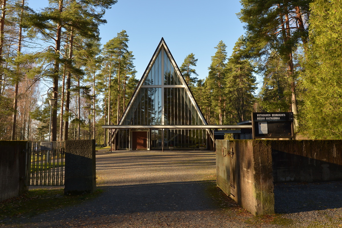 Kappelin lasinen, kolmion muotoinen julkisivu kuvassa sekä hieman ympäröivää puustoa ja pihaa.