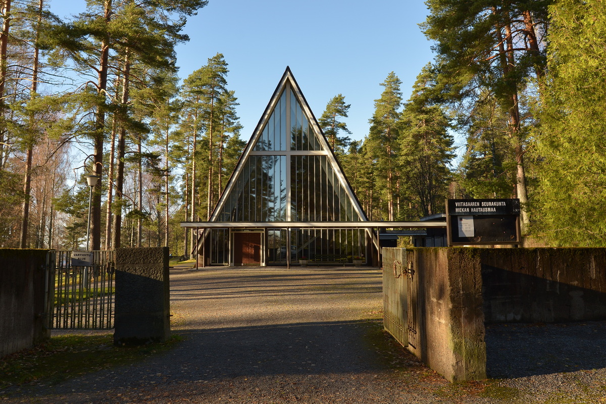 Siunauskappeli Kappelin lasinen, kolmion muotoinen julkisivu kuvassa sekä hieman ympäröivää puustoa ja pihaa.