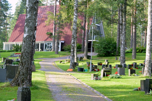Kappeli kuvattuna kesällä rannan suunnasta. Etualalla on käytävä ja hautoja, taustalla erottuu kappelin jyrkkä katto ja lasiseinää.